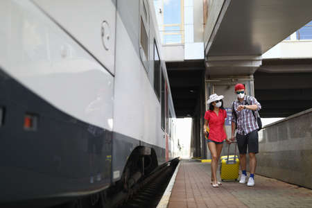 Man and woman in protective mask wait for arrival of train on platform at station with yellow suitcase.の写真素材