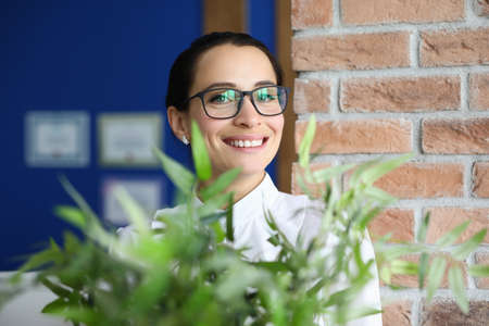 Happy office worker stand and smile at workplace with flower. Woman with glasses in office against background of brick wall and diplomas. HR department for professional staff recruitment.の写真素材