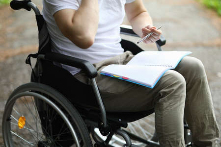 Man sit in wheelchair and take note in notebook in park. Disabled person keep diary and hold pen in his hand and notebook on his lap. Preparation of classes for rehabilitation after surgery.の写真素材