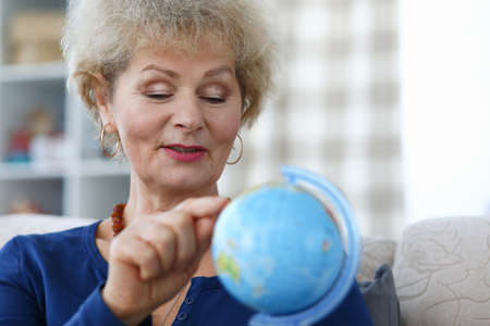 Elderly woman hold blue globe in her hand and point to continent with her index finger. Traveling around the world at retirement age. Exploration of world map. Travel agency carries out selection of tour.の写真素材