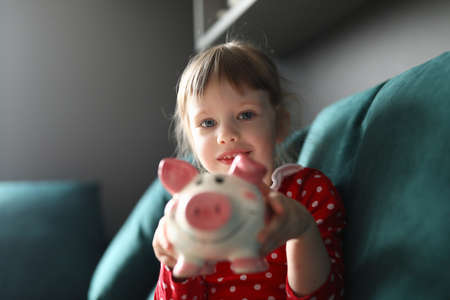 Girl in red dress with polka dots sit on couch and smile. Child hold pink piggy bank in her hands and show. Savings and accumulation of funds. Teaching thrift children.の写真素材