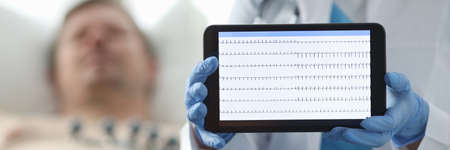 Doctor in clinic shows patients cardiogram tablet. Examination and diagnosis heart, cardiogram. Cardiogram is displayed on device screen. Discussion patients illness. Man lies on bed in clinicの写真素材