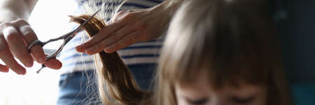 Close-up of mother holding strand of daughter hair and going to cut with metal scissors. Girl with...