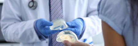 Close-up of professional doctor holding breast implant. Medical wearing uniform and red stethoscope. Patient at plastics appointment. Modern medicine and cosmetic surgeon conceptの写真素材