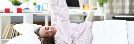 Portrait of sweet lady stretching in bed wearing comfy pyjamas. Lovely interior and cozy bed. Lazy day indoors under blanket. Morning routine and relaxation conceptの写真素材