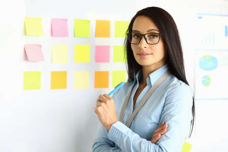 Businesswoman in blue shirt and glasses stand near blackboard with pen in her hand and smile....