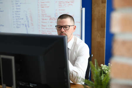 Serious man in glasses and shirt sit and look at computer monitor. In background is white board with...