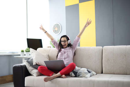 Woman on couch with a laptop happily raises her hands up and smiles. Online winnings on the internet...