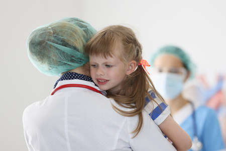 Doctor stands with his back and holds in his arms little frightened girl. Fear of child health care workers conceptの写真素材