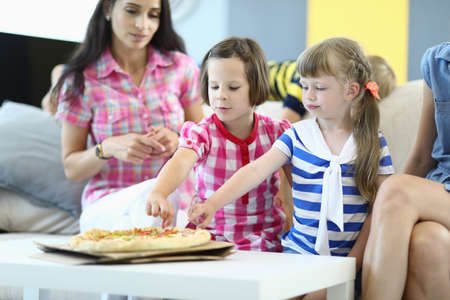 Little girls with mothers sit on couch and take slices of pizza. Childrens holiday conceptの写真素材