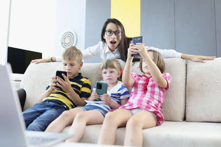 Three children are sitting on couch with smartphones in their hands, playing online games with woman...