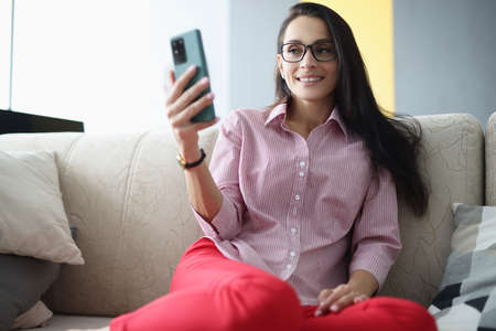 Woman with glasses sits on couch smiling and looks at smartphone screen. Remote work and freelance...