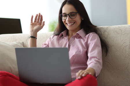Woman with glasses sits on couch smiles and greets with hand of the interlocutor in the laptop....