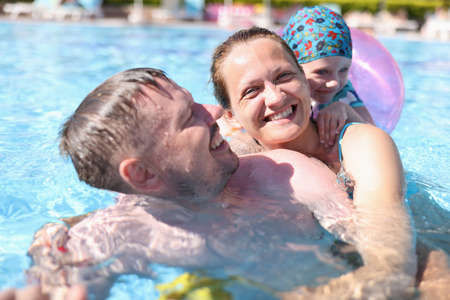 Happy family swim in pool. Beautiful woman hug man from behind in water. Girl with inflatable ring...