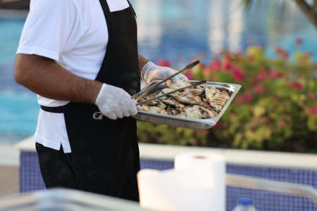 Man in black apron carries metal tray and spatula. Cook carries plate of grilled fried fish.の写真素材