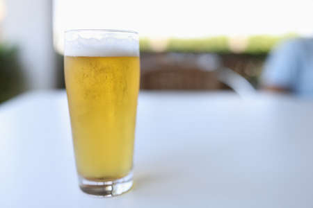 Glass is on white table outside. Glass of yellow cold beer with white foam is poured.の写真素材