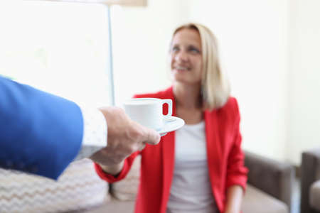 Man in blue jacket give white cup of coffee. Woman in red jacket sit on sofa and take cup and saucer.の写真素材