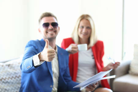 Man in blue jacket and sunglasses hold documents in his hand and show thumb up close-up with other...