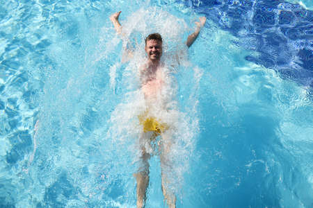 Happy man in yellow swimming trunks float on back in blue clear and transparent water. Splashes and waves from water fly in different directions.の写真素材