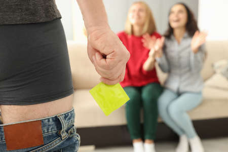 Man stands in shorts with his pants down and holds condom in front of two women. Relationship in...