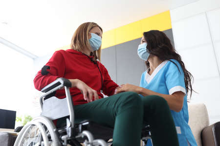Physiotherapist doctor holds hand of woman in wheelchair wearing medical masks. Psychological...