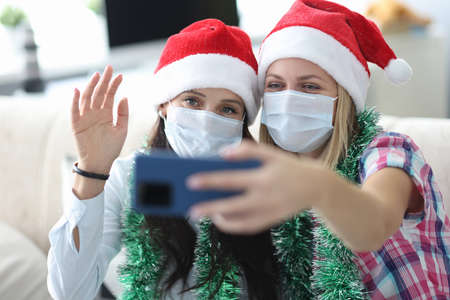 Two young women in santa hats and protective masks on faces are holding phone at home portrait. Online communication during covid 19 pandemic concept.の写真素材