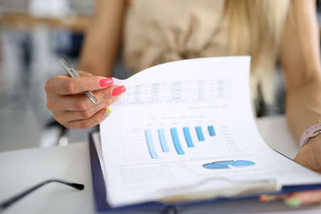 Female hand hold metal pen and leaf through documents. Woman sit at workplace and study charts.
