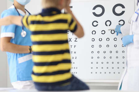 Boy in striped T-shirt sit on chair at ophthalmologist appointment and cover one eye with his hand. Doctor show metal pen to table with letters. Eyesight check.の写真素材