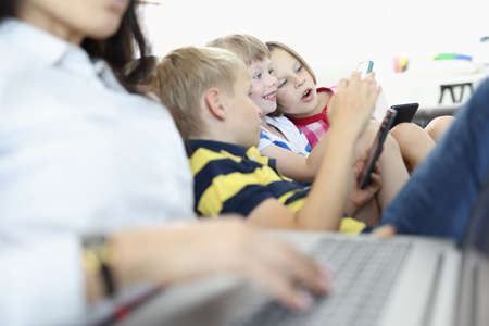 Three children sit side by side on couch and play on smartphones. Boy and girl look at phone screen in surprise. Woman is typing text on laptop next to children.の写真素材