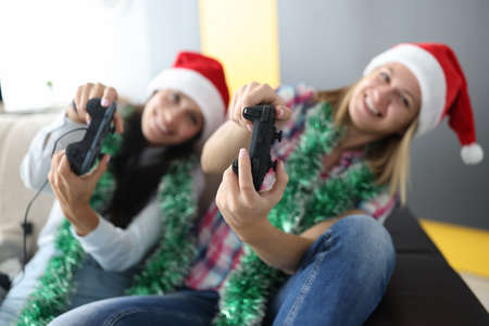 Two women in red santa claus hats with tinsel around their necks are sitting on couch with joysticks in hands and playing computer console. Entertainment for new years holidays at home concept.の写真素材