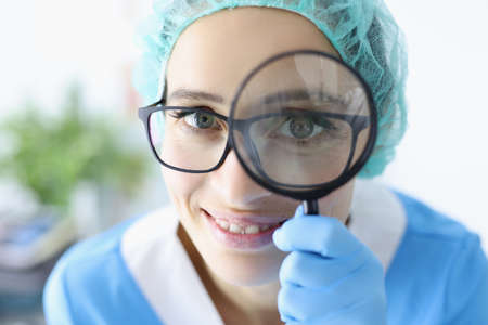 Young woman doctor in glasses and uniform holds magnifying glass near her eyes portrait. Dermological study of skin neoplasms concept.の写真素材