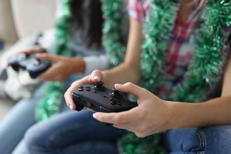 Two women with tinsel around their necks are holding joysticks close-up. Fun computer games new year...