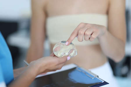 Woman patient with elastic bandage on her chest touches a silicone implant in doctors hand close-up....