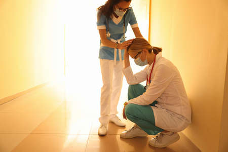 Female doctor in protective medical mask sits in corridor with her head lowered and her colleague supports her. Medical error in medicine conceptの写真素材