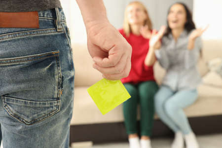 Two happy woman sit on couch and clap their hands. Man hold yellow condom in hand close up.