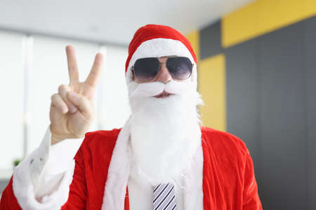 Man in santa claus costume with white beard and sunglasses holds his fingers in the sign of victory. Organization of surprises for children during New Year holidays conceptの写真素材