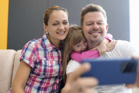 Family mom dad daughter sit in an embrace and photographed on smartphone. Family selfies for social media conceptの写真素材