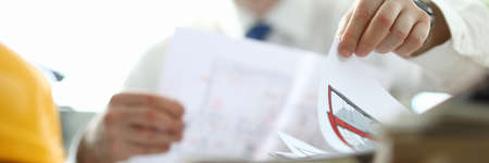 Close-up of businessman holding paper with house plan. Yellow helmet on working table. Smiling...