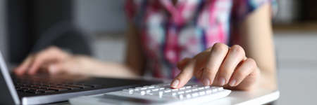 Close-up of young woman working on computer and typing on white calculator. Female accounting family budget on kitchen at home. Finance and saving up conceptの写真素材
