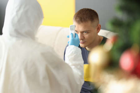 Doctor in protective suit measures temperature with non-contact thermometer to patient at home....