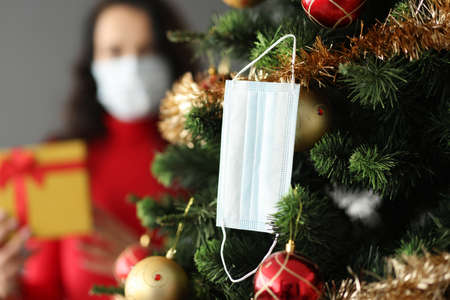 Medical protective mask weighs on tree. A woman in mask and gift stands behind. Celebrating New Year and Christmas in Coronavirus Pandemic Conceptの写真素材