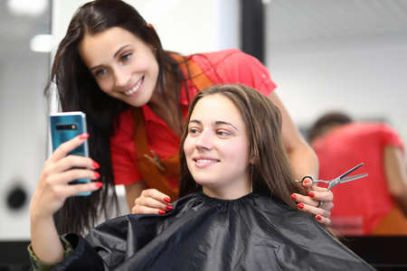 Master hairdresser with client look at smartphone screen in beauty salon. Selection of stylish and fashionable hairstyles for women conceptの写真素材