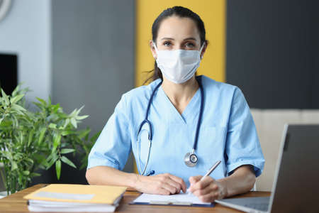 Doctor with protective medical mask siting at table in office and writing in documents. Medical...