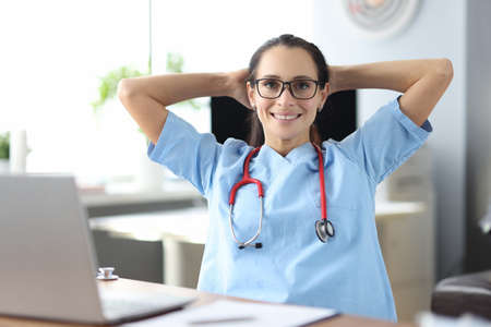 Young smiling doctor siting at table by laptop and holding head in clinic. Work break for medical staff conceptの写真素材