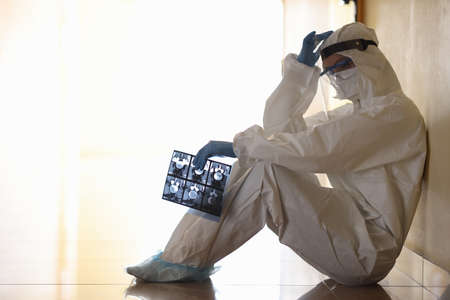 Tired doctor in protective anti-plague overalls siting in corridor of clinic. Emotional burnout in...