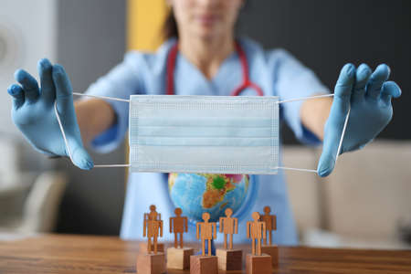 Doctor in rubber gloves holding protective medical face mask near globe and wooden men close-up....
