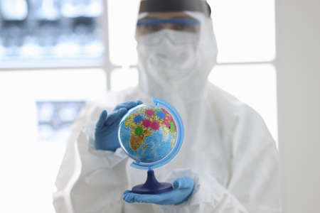 Doctor in protective suit and mask holds globe in his hands. Disease spread around the world conceptの写真素材