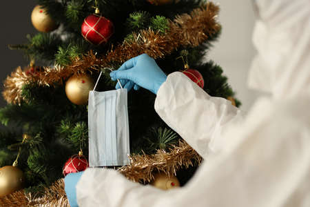 Doctor in protective antiplague suit hanging protective medical mask on Christmas tree. Safe new year celebration during covid19 pandemic conceptの写真素材