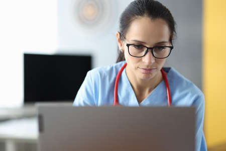 Young woman doctor in glasses siting at a laptop in clinic office. Online consultations during pandemic covid 19 conceptの写真素材