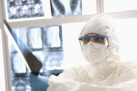 Tired doctor in protective suit and rubber gloves siting with x-ray in his hands in clinic....